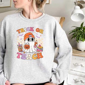 May include: Gray sweatshirt with a ghost wearing a hat and holding a book. The text "Trick or Teach" is printed on the front of the sweatshirt.