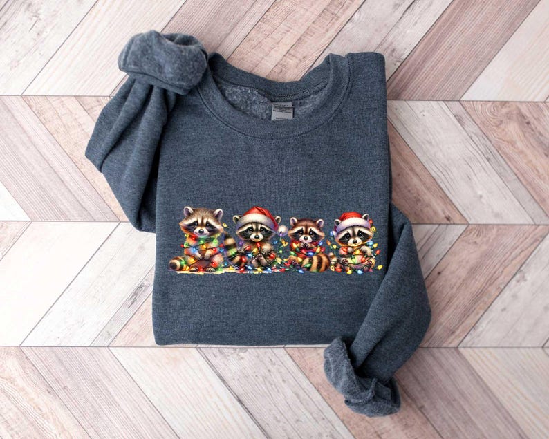 May include: A dark gray sweatshirt with a Christmas-themed design featuring four raccoons wearing Santa hats and holding strings of colorful lights.