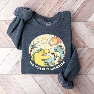 Peut inclure: Un sweat-shirt gris fonc&eacute; avec un imprim&eacute; graphique de deux dinosaures portant des lunettes de soleil. Les dinosaures sont debout sur une plage avec des palmiers et une com&egrave;te dans le ciel. Le texte "The vibe is in shambles" est imprim&eacute; sous les dinosaures.
