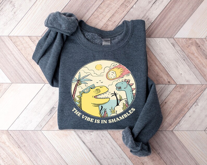 Peut inclure: Un sweat-shirt gris fonc&eacute; avec un imprim&eacute; graphique de deux dinosaures portant des lunettes de soleil. Les dinosaures sont debout sur une plage avec des palmiers et une com&egrave;te dans le ciel. Le texte "The vibe is in shambles" est imprim&eacute; sous les dinosaures.