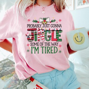 Peut inclure: Un sweat-shirt rose clair sur le th&egrave;me de No&euml;l avec le texte "PROBABLY JUST GONNA JINGLE SOME OF THE WAY I'M TIRED" dans une police festive. Le mot "JINGLE" est en lettres &agrave; carreaux et vertes. La personne tient une tasse blanche avec un smiley jaune.