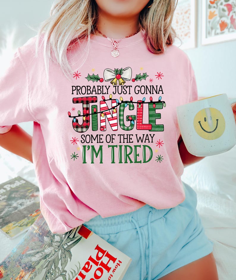 Peut inclure: Un sweat-shirt rose clair sur le th&egrave;me de No&euml;l avec le texte "PROBABLY JUST GONNA JINGLE SOME OF THE WAY I'M TIRED" dans une police festive. Le mot "JINGLE" est en lettres &agrave; carreaux et vertes. La personne tient une tasse blanche avec un smiley jaune.