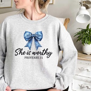 May include: A light gray sweatshirt featuring a blue bow graphic and the text "She is worthy" with "Proverbs 31" underneath. The sweatshirt is a crew neck style.