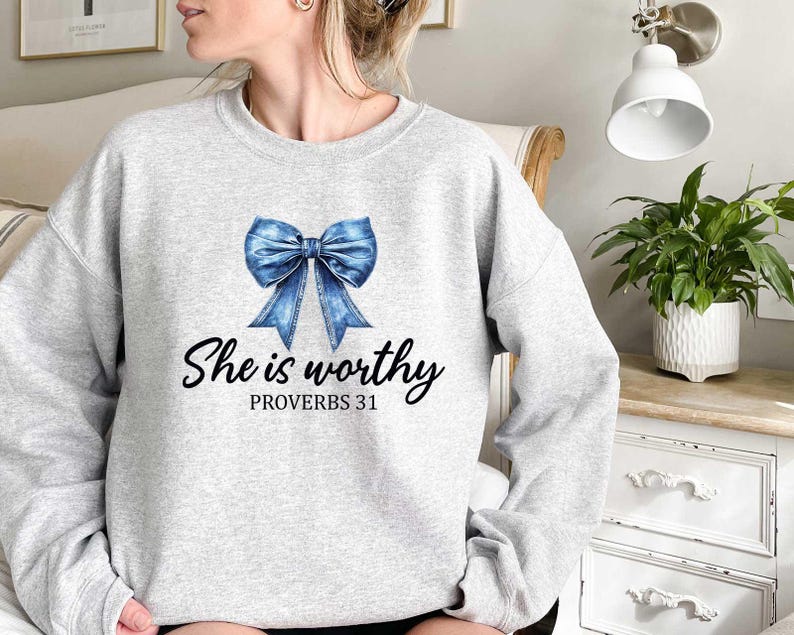 May include: A light gray sweatshirt featuring a blue bow graphic and the text "She is worthy" with "Proverbs 31" underneath. The sweatshirt is a crew neck style.