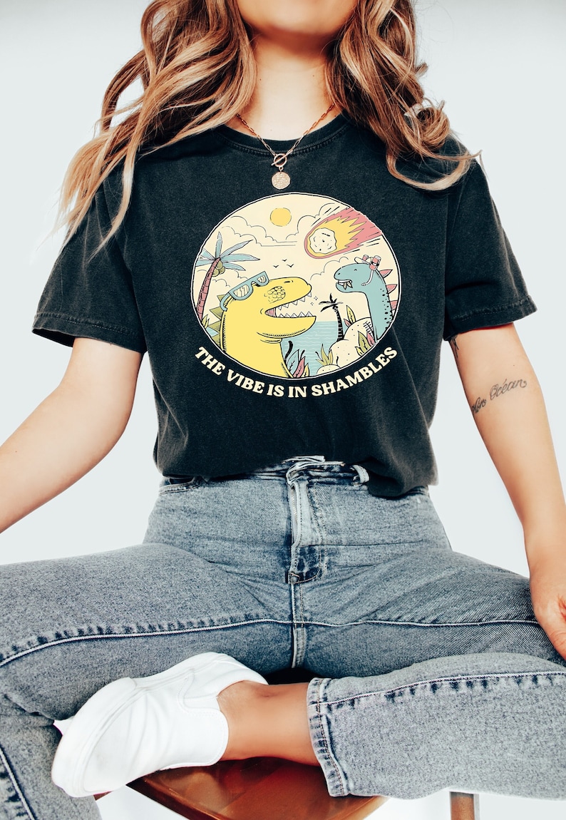 Peut inclure: Un t-shirt noir avec un motif de deux dinosaures dans un d&eacute;cor tropical. Le texte "The vibe is in shambles" est imprim&eacute; sous les dinosaures.
