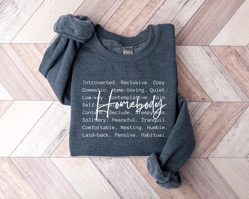 May include: A dark gray sweatshirt with the text "Introverted. Reclusive. Cozy. Domestic. Home-loving. Quiet. Low-key. Contemplative. Calm. Self Homebody Content. Seclude. Sleepyness. Solitary. Peaceful. Tranquil. Comfortable. Nesting. Humble. Laid-back. Pensive. Habitual" printed on the front.