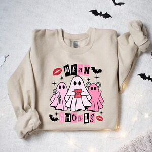 May include: A beige sweatshirt with a graphic print of three pink ghosts with the text "Mean Ghouls" in black and pink letters. The ghosts are holding items like a key and a heart.