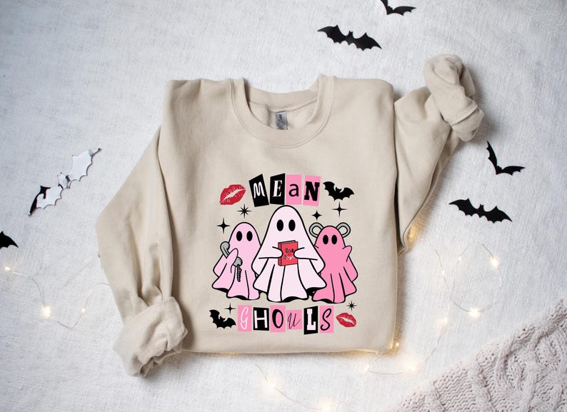 May include: A beige sweatshirt with a graphic print of three pink ghosts with the text "Mean Ghouls" in black and pink letters. The ghosts are holding items like a key and a heart.