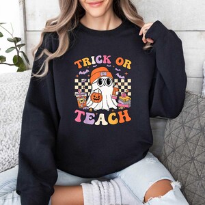 May include: Black sweatshirt with a graphic of a ghost wearing glasses and a graduation cap. The text "Trick or Teach" is printed in rainbow colors.