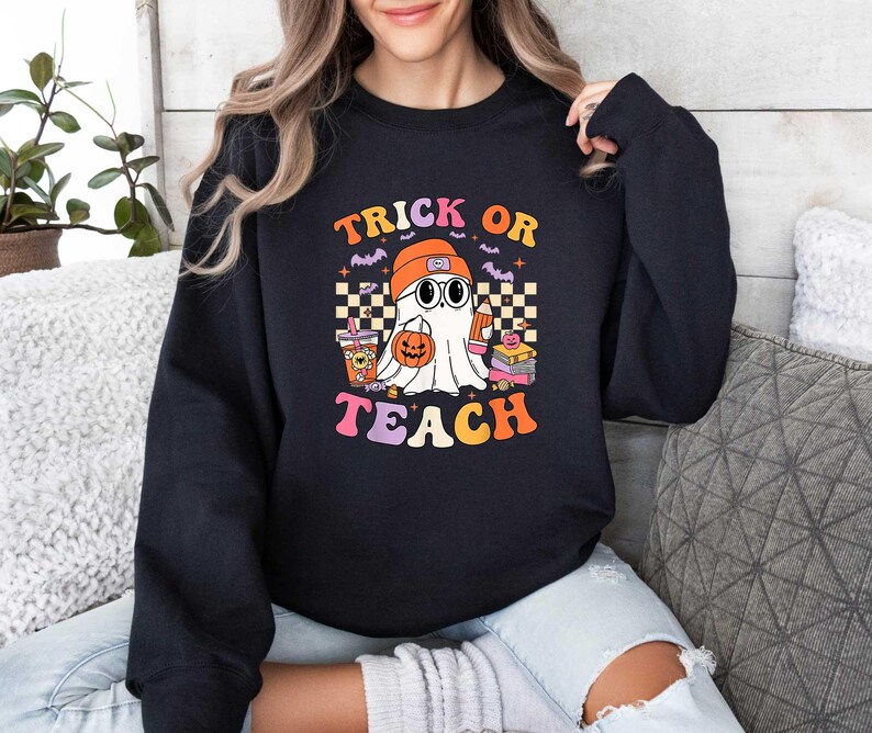 May include: Black sweatshirt with a graphic of a ghost wearing glasses and a graduation cap. The text "Trick or Teach" is printed in rainbow colors.