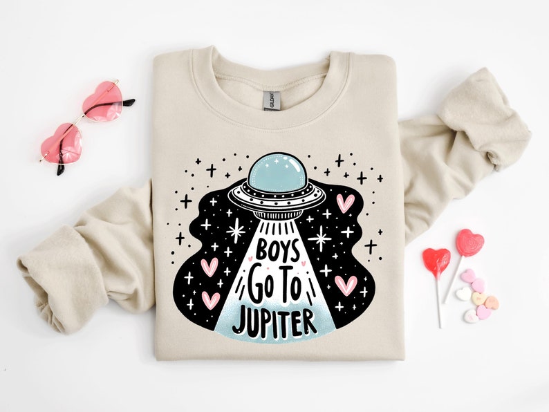 以下が含まれることがあります： ベージュのトレーナーに、UFOと「Boys Go To Jupiter」の文字、ピンクのハートが描かれた黒と白のグラフィックがプリントされています。