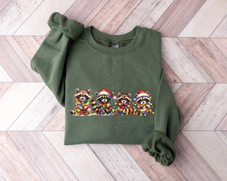May include: A green sweatshirt with a Christmas-themed design featuring four raccoons wearing Santa hats and holding Christmas lights.