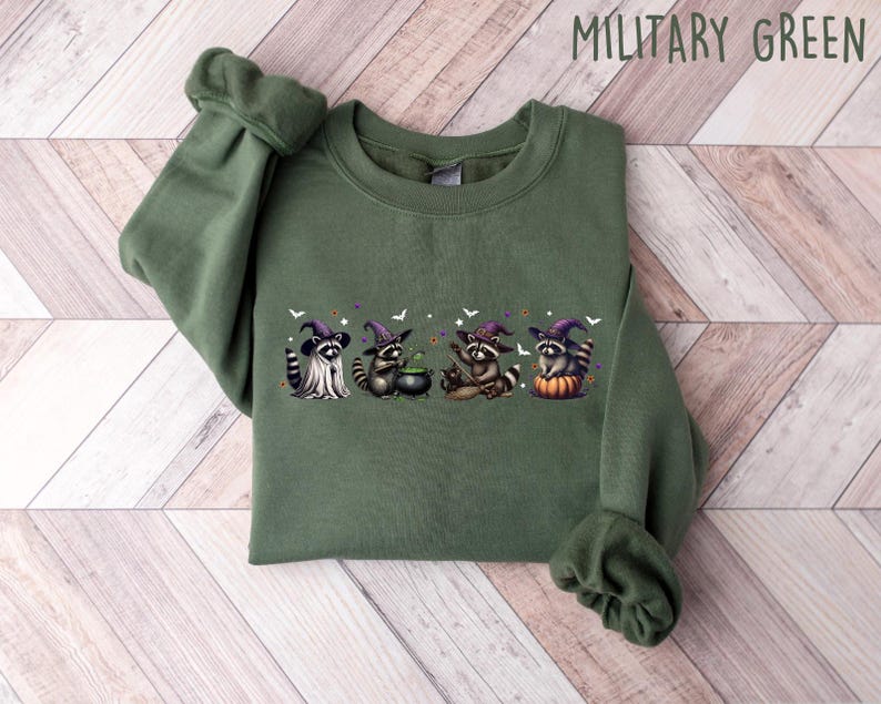 May include: Military green sweatshirt with a Halloween graphic. The design shows four raccoons in witch hats and costumes, with a cauldron and bats. The sweatshirt is made of a soft material and has ribbed cuffs and a crew neck. The text "MILITARY GREEN" is visible.