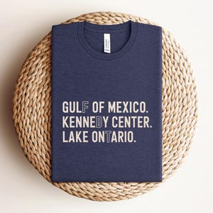 May include: A navy blue crew neck t-shirt with light beige text printed on the front. The text reads: GULF OF MEXICO. KENNEDY CENTER. LAKE ONTARIO. The shirt features a white brand label inside the collar and has a short-sleeved design.