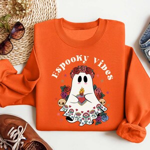 May include: Orange sweatshirt with a white ghost wearing a floral crown and holding a heart. The text "Spooky Vibes" is printed on the sweatshirt.