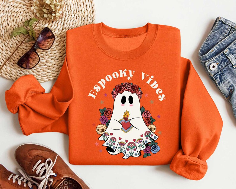May include: Orange sweatshirt with a white ghost wearing a floral crown and holding a heart. The text "Spooky Vibes" is printed on the sweatshirt.