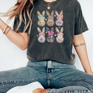 May include: A dark gray t-shirt with a graphic print of six colorful Easter bunnies. The bunnies are arranged in a grid pattern and each has a different floral design. The shirt has a relaxed fit and a crew neckline.