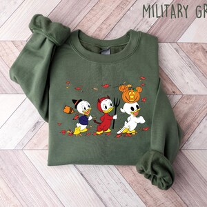 May include: A green sweatshirt featuring a graphic print of three cartoon ducks dressed in Halloween costumes. The ducks are wearing a witch hat, devil horns, and a ghost costume. The text "MILITARY GREEN" is printed in the top right corner of the image.