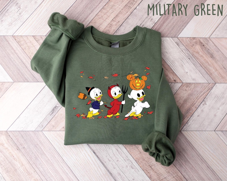 May include: A green sweatshirt featuring a graphic print of three cartoon ducks dressed in Halloween costumes. The ducks are wearing a witch hat, devil horns, and a ghost costume. The text "MILITARY GREEN" is printed in the top right corner of the image.
