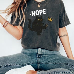 May include: Dark grey t-shirt with the word "-NOPE" in white. A black cat with yellow eyes kicks a gold crown. The shirt is a casual style.
