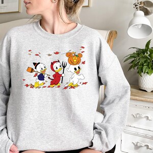 May include: A grey sweatshirt featuring a graphic of three cartoon ducks dressed in Halloween costumes. The ducks are walking through a field of autumn leaves. The duck in the middle is wearing a red devil costume, the duck on the left is wearing a witch's hat, and the duck on the right is wearing a ghost costume.