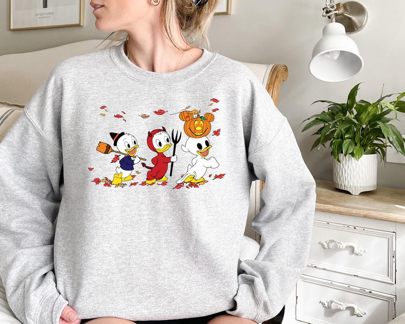 May include: A grey sweatshirt featuring a graphic of three cartoon ducks dressed in Halloween costumes. The ducks are walking through a field of autumn leaves. The duck in the middle is wearing a red devil costume, the duck on the left is wearing a witch's hat, and the duck on the right is wearing a ghost costume.