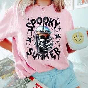 May include: Pink t-shirt with a graphic of a skeleton hand holding a black iced coffee cup with the text "Spooky Summer" in dripping black letters and stars.