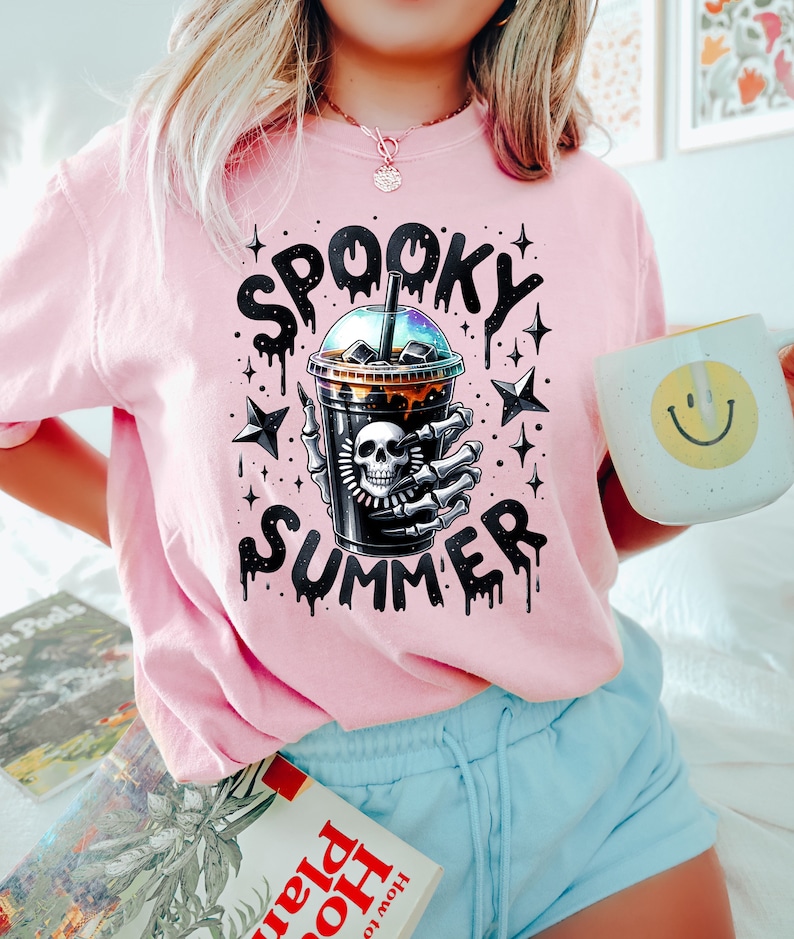 May include: Pink t-shirt with a graphic of a skeleton hand holding a black iced coffee cup with the text "Spooky Summer" in dripping black letters and stars.