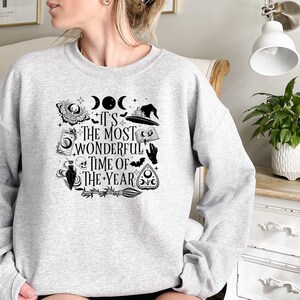 Pu&ograve; includere: Una felpa grigia con una stampa grafica in bianco e nero di un cappello da strega, una luna crescente, un teschio, una mano e un pentagramma. Il testo sulla felpa recita "It's the most wonderful time of the year."