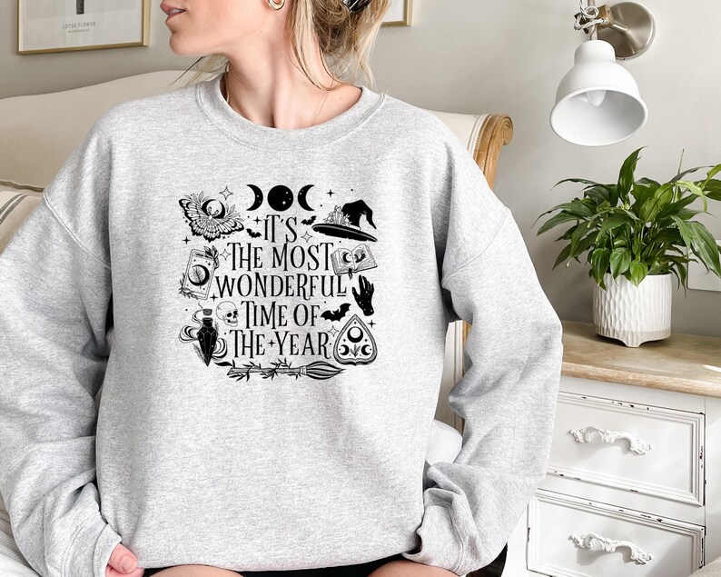 Pu&ograve; includere: Una felpa grigia con una stampa grafica in bianco e nero di un cappello da strega, una luna crescente, un teschio, una mano e un pentagramma. Il testo sulla felpa recita "It's the most wonderful time of the year."