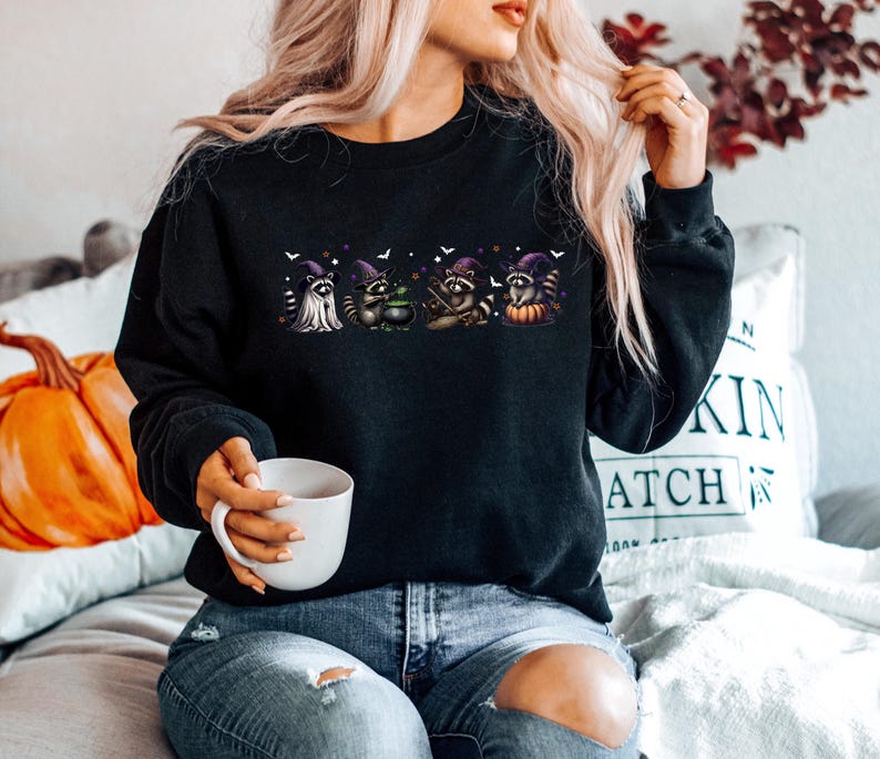 May include: Black sweatshirt with a Halloween design. The graphic shows four raccoons in witch costumes, a ghost, and a pumpkin. The raccoons wear purple witch hats, with bats and stars around them. The sweatshirt is a crew neck style.