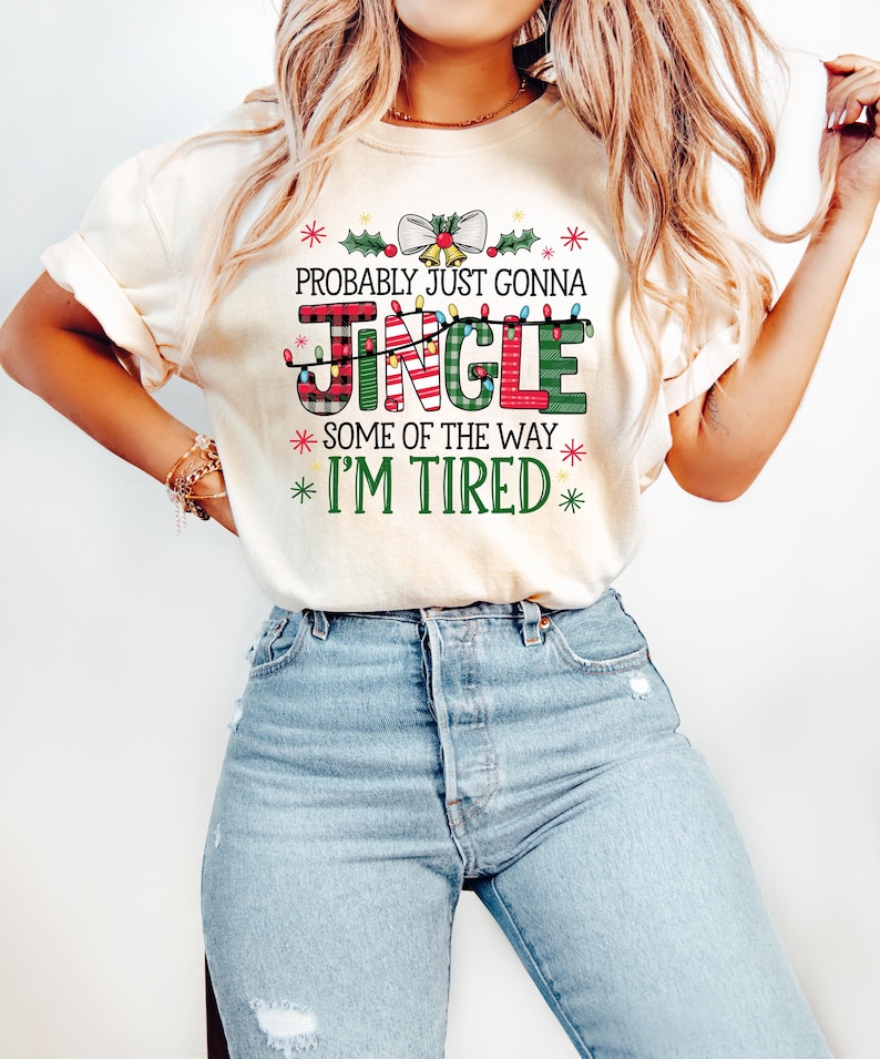 Peut inclure: T-shirt cr&egrave;me avec un motif sur le th&egrave;me de No&euml;l. Le design comprend les mots "Probably Just Gonna Jingle Some of the Way I'm Tired" dans un m&eacute;lange de polices et de couleurs, avec des accents festifs comme du houx et des lumi&egrave;res.
