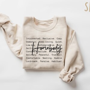 May include: A beige sweatshirt with the word "Homebody" written in black cursive lettering. The sweatshirt has a list of words describing a homebody personality, such as "introverted", "reclusive", "cozy", "domestic", "quiet", "calm", "peaceful", "tranquil", "comfortable", "humble", "laid-back", "pensive", and "habitual".