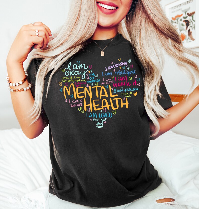 Op de afbeelding: Zwart T-shirt met een hartvormig ontwerp met de woorden "Geestelijke gezondheid" in geel en andere positieve affirmaties in verschillende kleuren. De tekst bevat zinnen als "Ik ben ok&eacute;", "Ik ben sterk", "Ik ben geliefd" en "Ik kan dit".