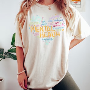 Op de afbeelding: Een cr&egrave;mekleurig T-shirt met een kleurrijk grafisch ontwerp met de tekst "Ik ben sterk! Ik verdien liefde. Ik ben een krijger. Ik ben zo sterk! Ik ben niet alleen. Ik ben intelligent. Ik ben het waard. Ik ben kostbaar. Geestelijke gezondheid. Ik ben geliefd."