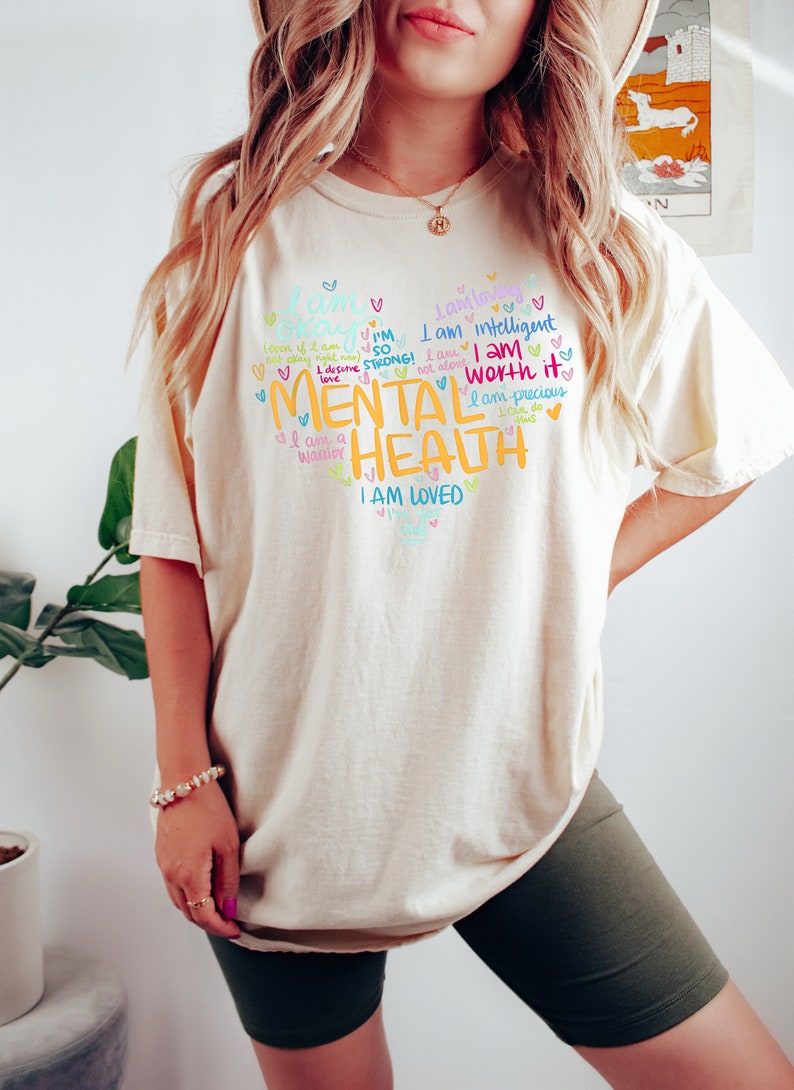 Op de afbeelding: Een cr&egrave;mekleurig T-shirt met een kleurrijk grafisch ontwerp met de tekst "Ik ben sterk! Ik verdien liefde. Ik ben een krijger. Ik ben zo sterk! Ik ben niet alleen. Ik ben intelligent. Ik ben het waard. Ik ben kostbaar. Geestelijke gezondheid. Ik ben geliefd."
