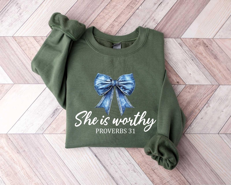 May include: An olive green sweatshirt featuring a denim-look bow graphic. The text "She is worthy" and "PROVERBS 31" are printed in white below the bow. The sweatshirt has long sleeves with ribbed cuffs.