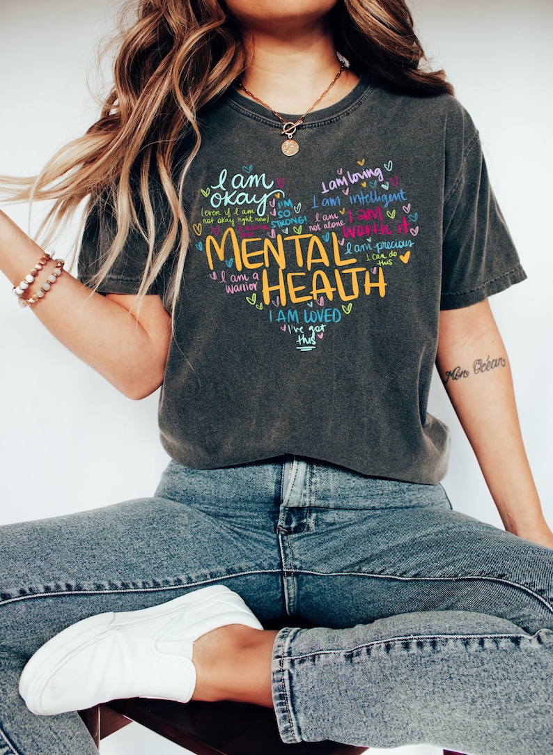 Op de afbeelding: Een donkergrijs T-shirt met een grote hartvormige afbeelding met de woorden "Geestelijke gezondheid" in geel. Het hart is gevuld met kleurrijke handgeschreven affirmaties, waaronder "Ik ben ok&eacute;", "Ik ben sterk", "Ik ben het waard", "Ik ben geliefd" en "Ik kan dit".
