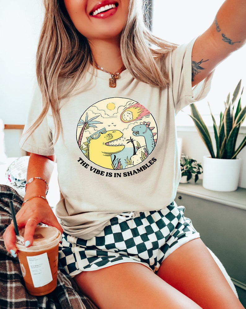 Peut inclure: Un t-shirt beige avec un motif de deux dinosaures dans un d&eacute;cor tropical. Le texte "The vibe is in shambles" est imprim&eacute; sous le motif.