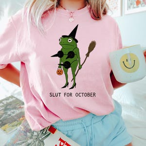 May include: Pink crew neck t-shirt featuring a green frog dressed as a witch with a black hat, holding a broom and a jack-o'-lantern. The text "SLUT FOR OCTOBER" is printed below the frog.