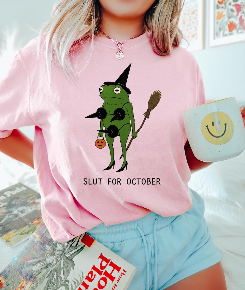 May include: Pink crew neck t-shirt featuring a green frog dressed as a witch with a black hat, holding a broom and a jack-o'-lantern. The text "SLUT FOR OCTOBER" is printed below the frog.