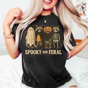 May include: Black t-shirt with a Halloween-themed graphic. The design features a ghost, two raccoons, and a pumpkin-headed raccoon. The text "SPOOKY AND FERAL" is printed below the characters. The shirt is worn by a person.