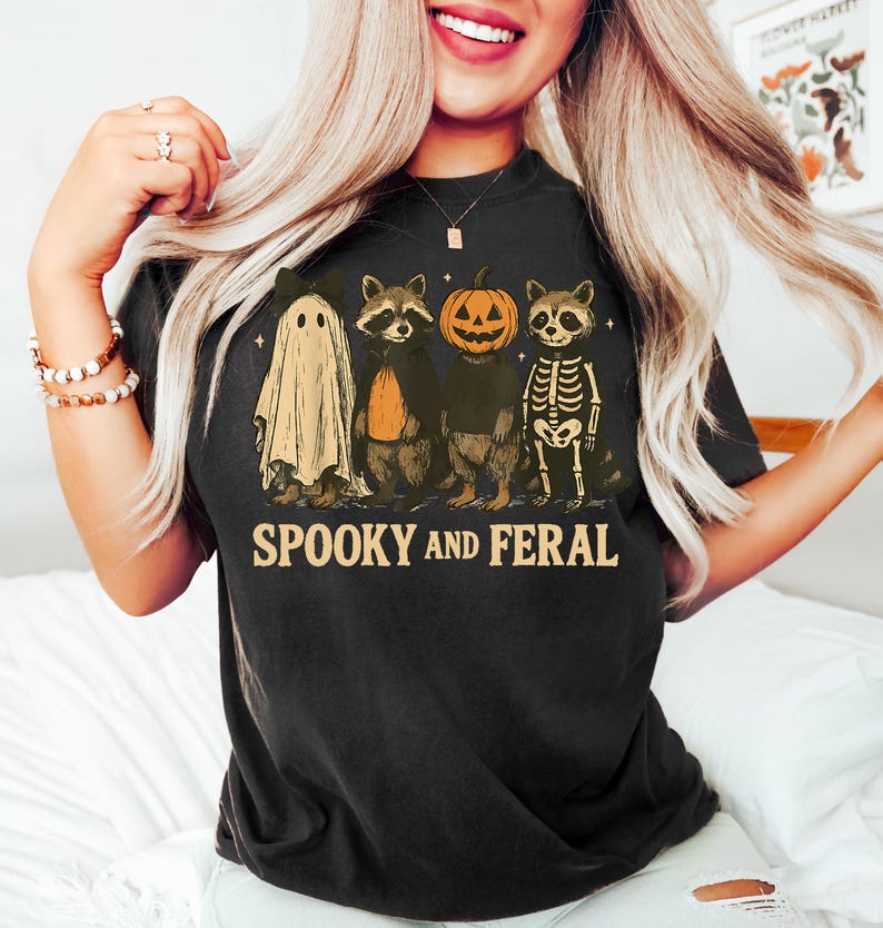 May include: Black t-shirt with a Halloween-themed graphic. The design features a ghost, two raccoons, and a pumpkin-headed raccoon. The text "SPOOKY AND FERAL" is printed below the characters. The shirt is worn by a person.