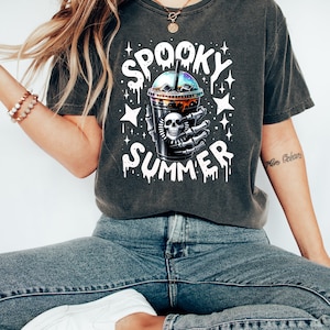 May include: A dark gray t-shirt with a white graphic of a skull holding a spooky summer drink. The text "Spooky Summer" is dripping down the sides of the graphic.