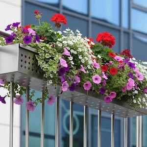Könnte beinhalten: Ein Balkon-Blumenkasten aus Edelstahl, überquellend mit leuchtenden Blumen. Die Blüten umfassen rote Geranien, lila Petunien und weißen Steinklee. Der Pflanzkasten ist an einem Metallgeländer befestigt.