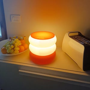 Puede incluir: Una lámpara de mesa de varios niveles de color blanco y naranja con una base blanca y una parte superior naranja. La lámpara está sobre una encimera blanca con un cuenco de fruta y una tostadora blanca.