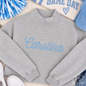 May include: A heather gray mock neck sweatshirt featuring the word Carolina embroidered in light blue cursive script across the chest. The garment has long sleeves with ribbed cuffs and a ribbed mock neckline, providing a clean and structured look.