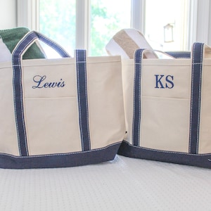 May include: Two canvas tote bags with navy blue trim and handles. One bag has "Lewis" embroidered in navy blue, the other has "KS". Both bags have a cream-colored body and a navy blue base. The bags are sitting on a white surface.