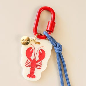 May include: A lobster-shaped keychain with a red lobster design on a white background. It features a red carabiner clip, a gold-colored charm, and a blue cord. The keychain is designed for attaching to keys or bags.