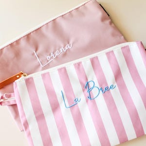 May include: Two zippered pouches. One is solid dusty pink with the name Lorena embroidered in white thread. The other is white with vertical pink stripes and the name La Bree embroidered in light blue thread. Both have white zippers.